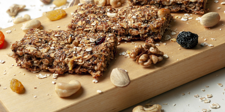 bar with granola, dried fruits and nuts on a light wooden board with dried fruits, nuts and cereals scattered nearby, close-up.