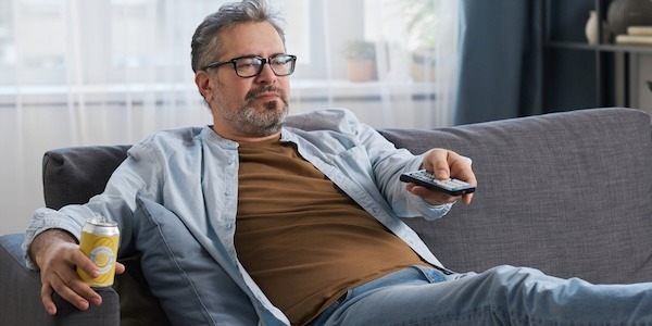 Mature man with drink resting on sofa and switching channels with remote control while watching TV at home